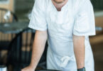 Male chef in a white uniform leaning on a striped chair at a table in a restaurant setting.