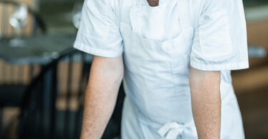 Male chef in a white uniform leaning on a striped chair at a table in a restaurant setting.