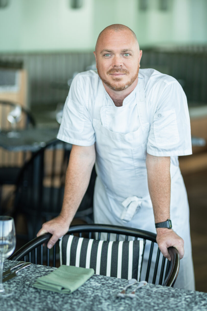 Male chef in a white uniform leaning on a striped chair at a table in a restaurant setting.