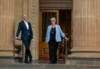 Two adults in business attire descend stone steps from a grand columned building, smiling at the camera.