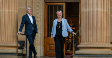 Two adults in business attire descend stone steps from a grand columned building, smiling at the camera.