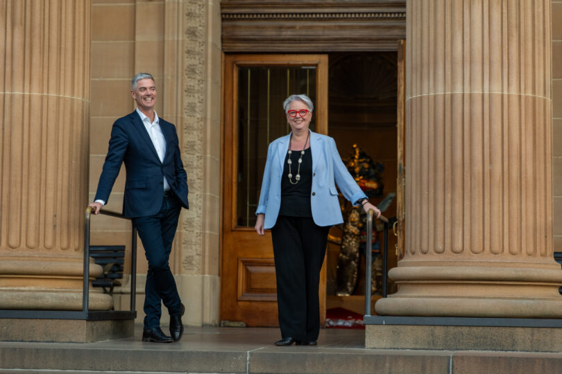 Two adults in business attire descend stone steps from a grand columned building, smiling at the camera.
