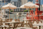 Outdoor cafe seating with beige umbrellas and metal chairs overlooking a marina and city skyline.