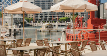 Outdoor cafe seating with beige umbrellas and metal chairs overlooking a marina and city skyline.