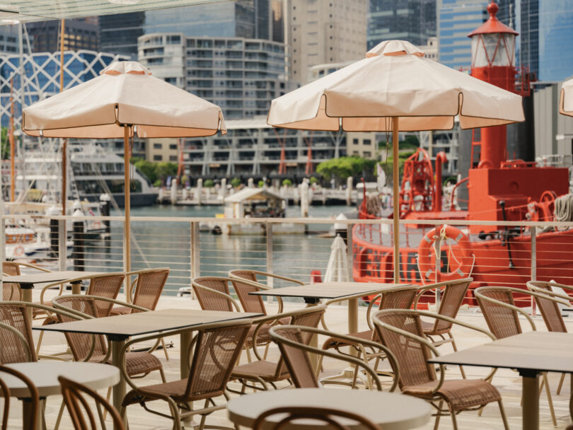 Outdoor cafe seating with beige umbrellas and metal chairs overlooking a marina and city skyline.