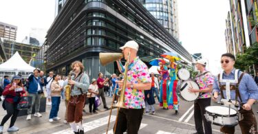 Bright street parade with musicians in colorful outfits playing brass instruments amid a crowd in an urban plaza behind modern buildings, sunny day.
