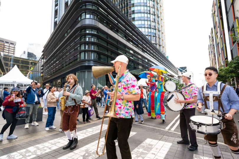 Bright street parade with musicians in colorful outfits playing brass instruments amid a crowd in an urban plaza behind modern buildings, sunny day.