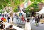 Crowded outdoor market with white stalls, shoppers, and a tall blue cart carrying a performer with a pink umbrella.