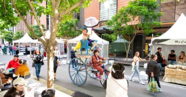 Crowded outdoor market with white stalls, shoppers, and a tall blue cart carrying a performer with a pink umbrella.