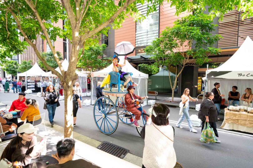 Crowded outdoor market with white stalls, shoppers, and a tall blue cart carrying a performer with a pink umbrella.