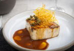 Seared rectangular piece of protein with a brown glaze, crispy potato strings on top, and green peppercorns on a white plate.