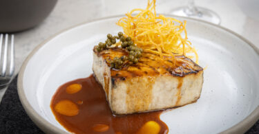 Seared rectangular piece of protein with a brown glaze, crispy potato strings on top, and green peppercorns on a white plate.
