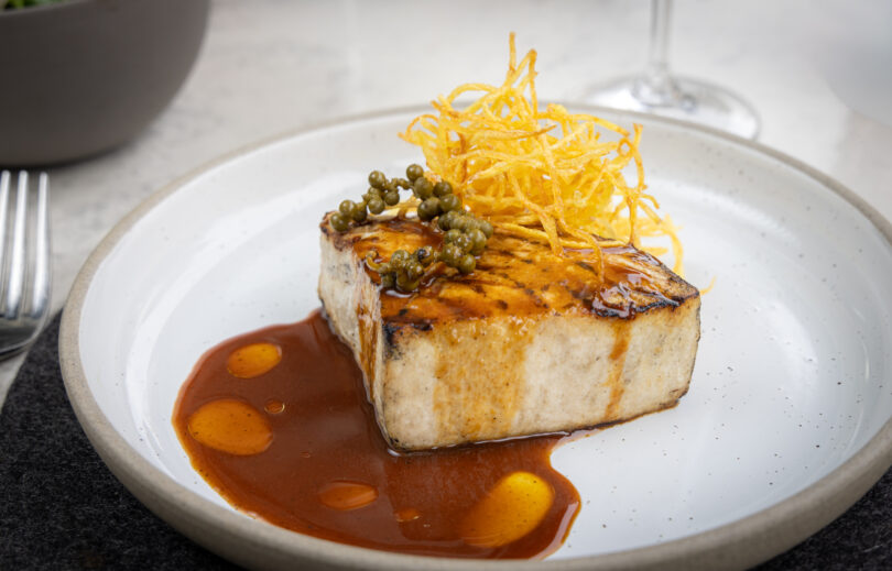 Seared rectangular piece of protein with a brown glaze, crispy potato strings on top, and green peppercorns on a white plate.