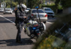 Australian NSW Police Force motorcycle officer in protective gear standing beside his parked police motorbike on a residential street with blue emergency lights flashing in the back.