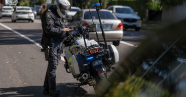 Australian NSW Police Force motorcycle officer in protective gear standing beside his parked police motorbike on a residential street with blue emergency lights flashing in the back.