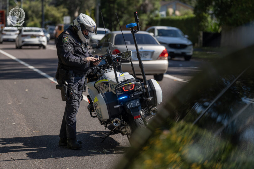 Australian NSW Police Force motorcycle officer in protective gear standing beside his parked police motorbike on a residential street with blue emergency lights flashing in the back.