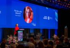 Speaker at podium in red dress presenting on a blue conference stage with giant screen reading 'The Hon. Julia Gillard AC' and 'Keynote Speaker'
