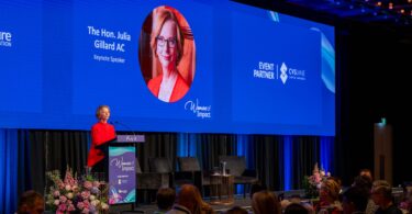 Speaker at podium in red dress presenting on a blue conference stage with giant screen reading 'The Hon. Julia Gillard AC' and 'Keynote Speaker'