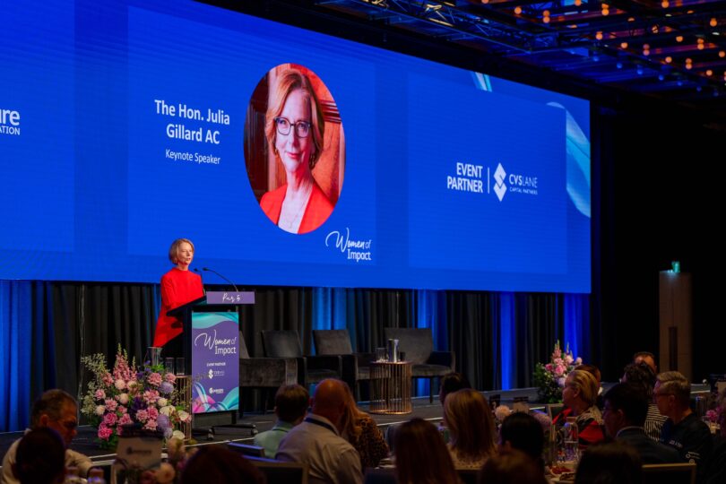 Speaker at podium in red dress presenting on a blue conference stage with giant screen reading 'The Hon. Julia Gillard AC' and 'Keynote Speaker'