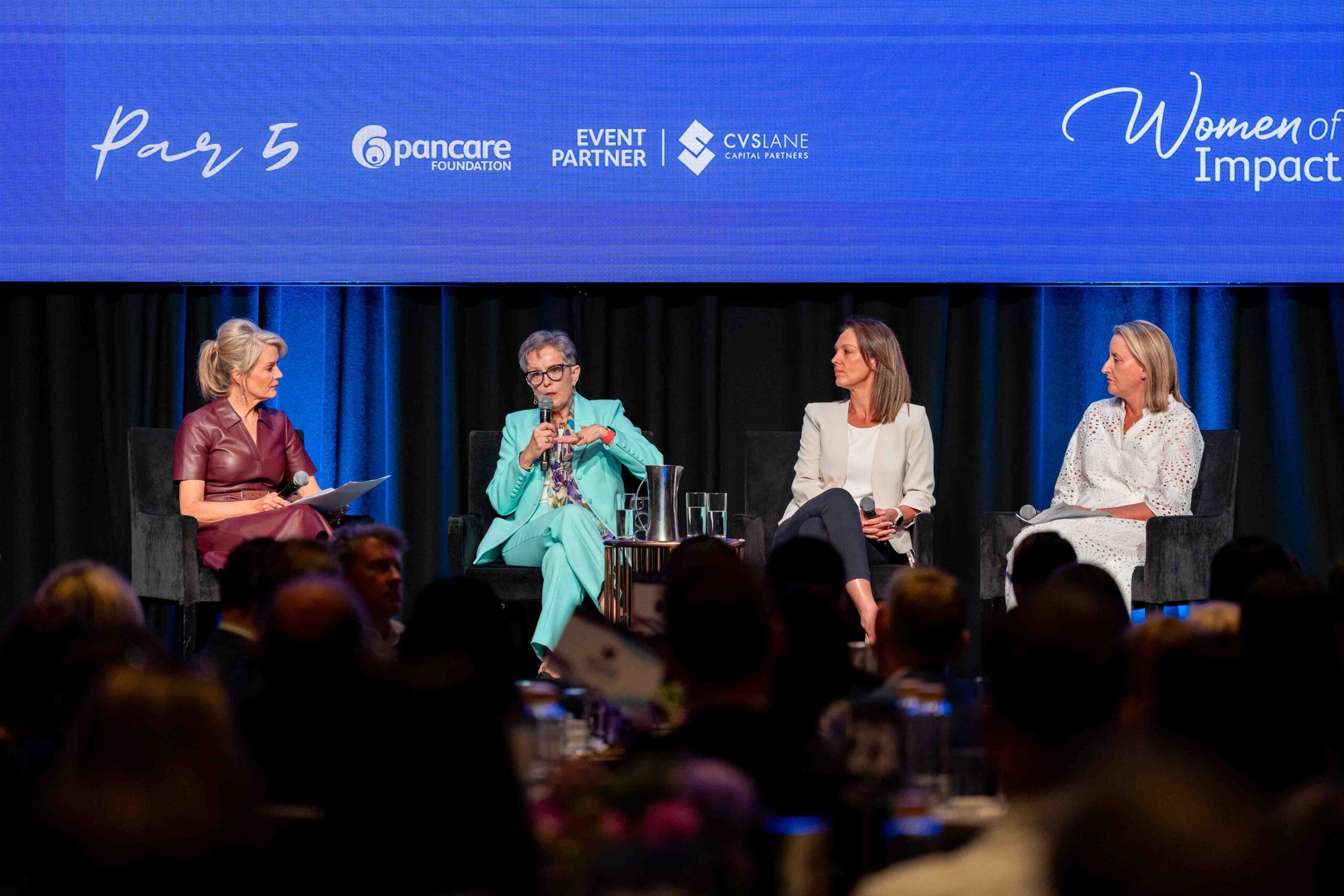 Four women panelists seated on a stage under a blue backdrop, one speaking into a microphone.