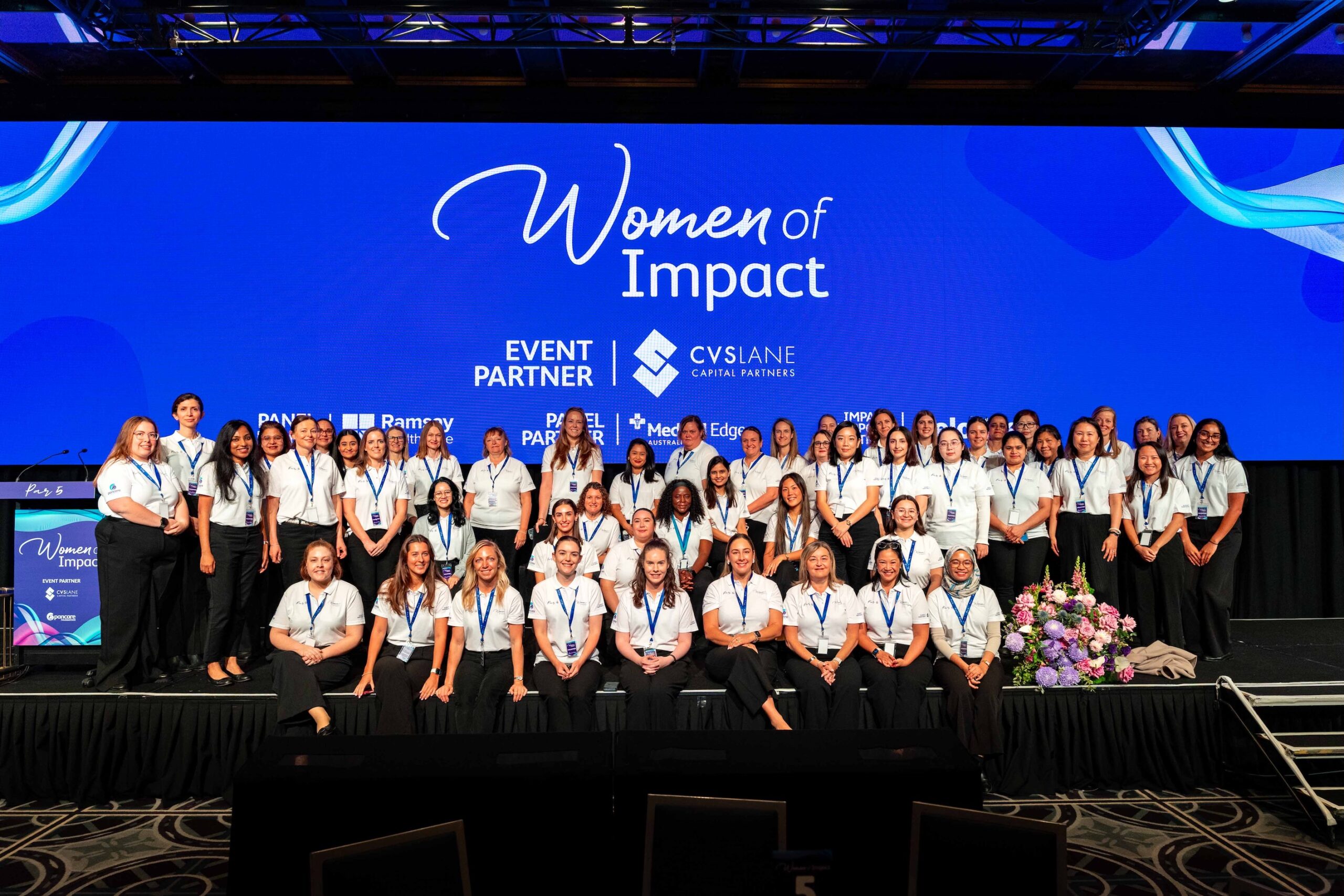Group of women in white shirts with lanyards posing on a stage at the Women of Impact event with a blue backdrop behind them.