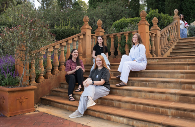 Five friends pose for a photo on a terracotta-colored garden staircase with ornate railings, smiling at the camera.