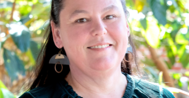 Portrait of a smiling woman with dark hair wearing a teal blouse and geometric earrings, outdoors with green plants in the background.