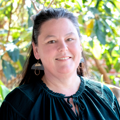 Portrait of a smiling woman with dark hair wearing a teal blouse and geometric earrings, outdoors with green plants in the background.