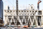 Construction site of a modern building with a white lattice framework and tall cranes over a waterway.
