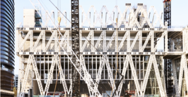 Construction site of a modern building with a white lattice framework and tall cranes over a waterway.
