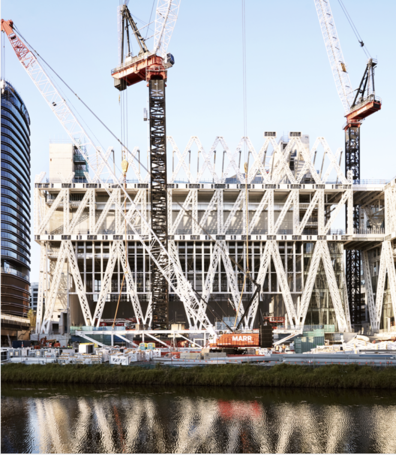 Construction site of a modern building with a white lattice framework and tall cranes over a waterway.